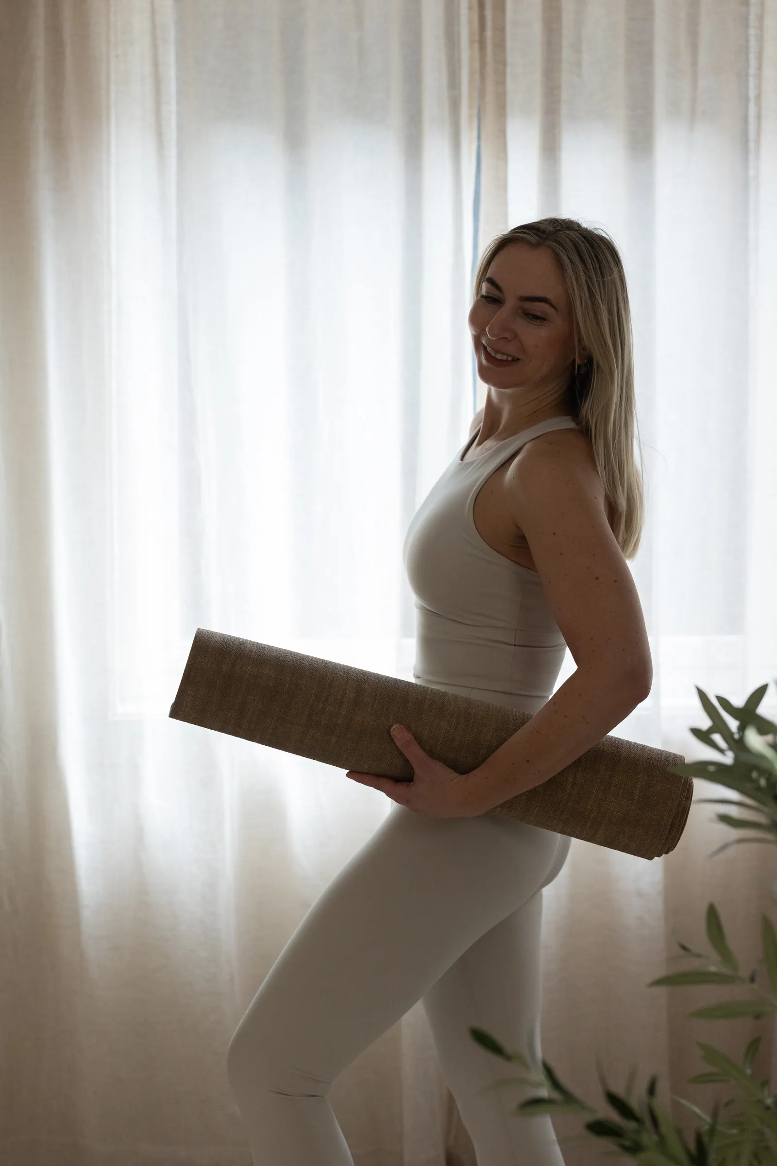 Larysa står smilende med yogamatte foran lyse gardiner i studioet