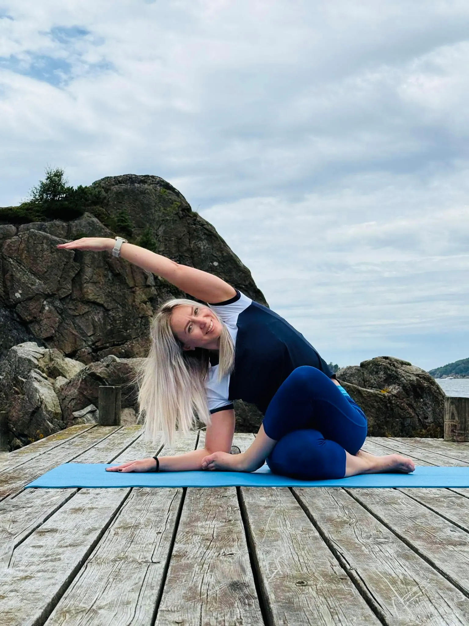 Sittende sidestrekk på blå yogamatte på en trebrygge ved sjøen, med fjell og åpen himmel i bakgrunnen
