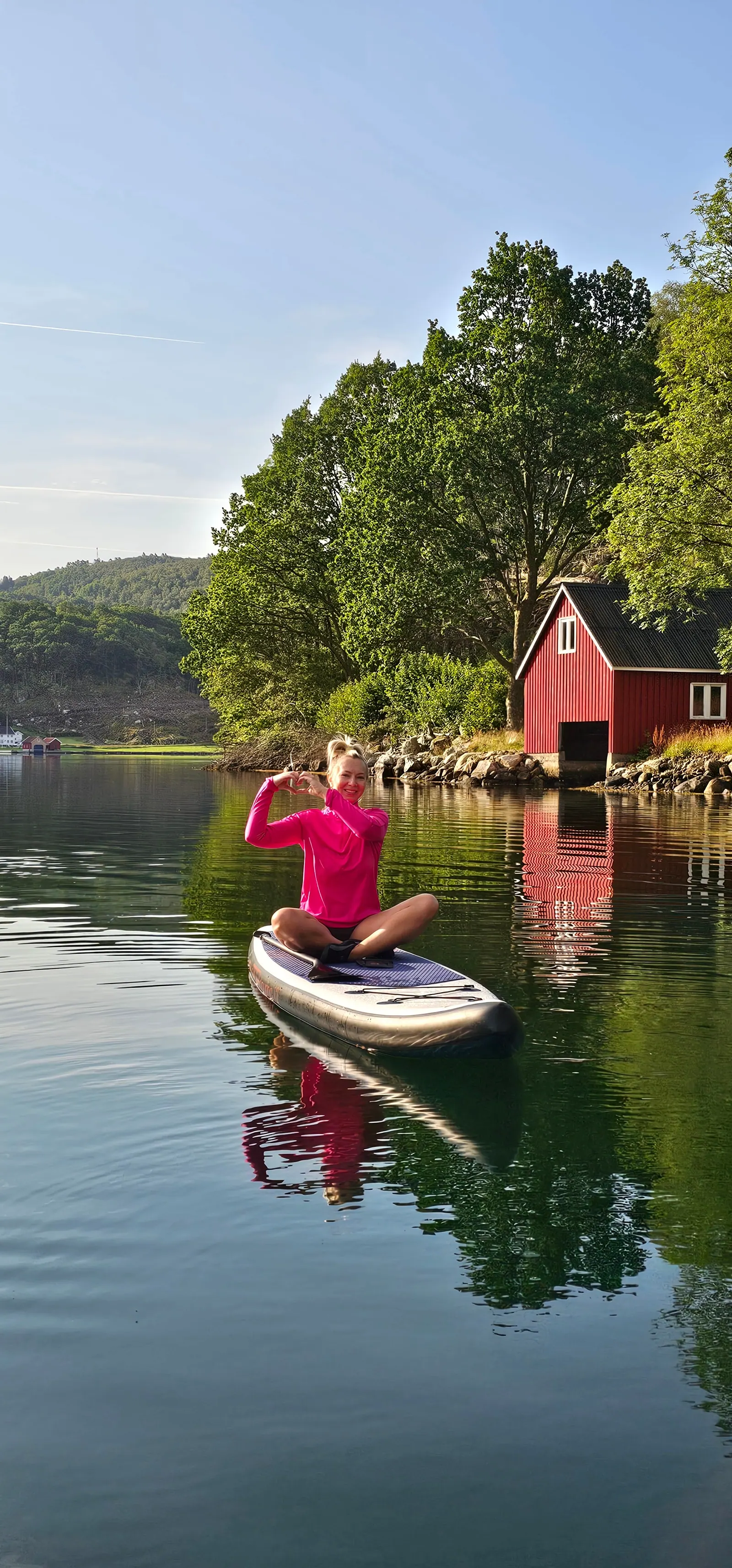 Sittende på SUP-brett i stille fjord ved et rødt sjøhus