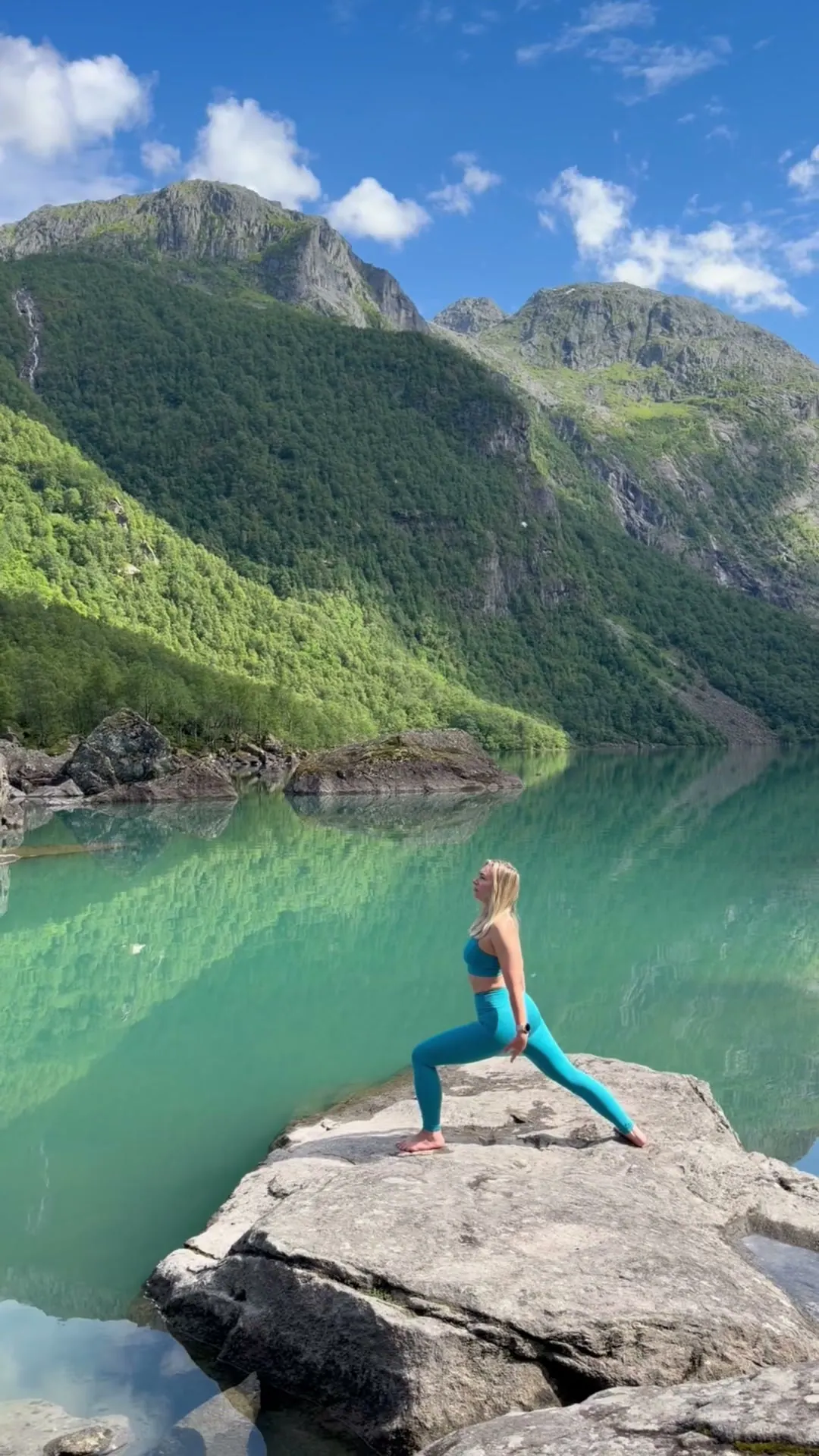 Yogastilling på en stein ved en smaragdgrønn innsjø, med grønne fjell som speiler seg i vannet